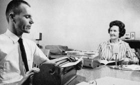 Man sitting at typewriter on left, woman, seated, on right.