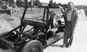 One man sitting in experimental automobile frame, another standing next to it